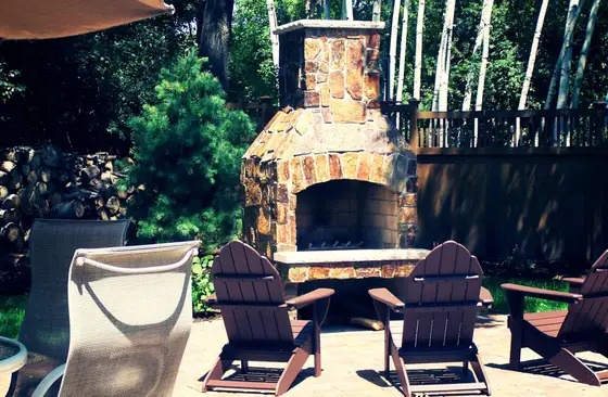 Stacked-stone outdoor fireplace with wood Adirondack chairs on a paver patio surrounded by evergreens