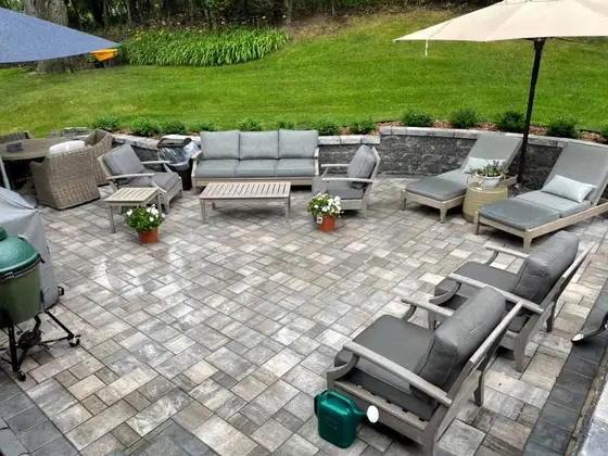 Paver patio with a stone seating wall, cushioned outdoor furniture, and grill overlooking a green backyard