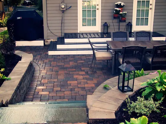 Tumbled paver patio with a low retaining wall border, grill, and outdoor dining set off a home's deck
