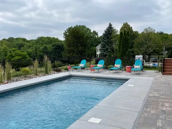 Inground pool with a paver deck, lounge chairs, and black fencing along a wooded backyard