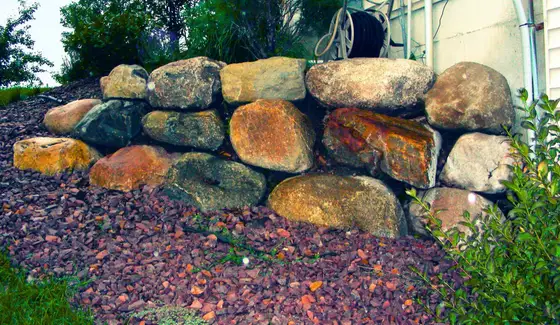 Natural boulder retaining wall bordered by red rock mulch along the side of a house