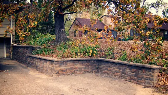 Curved segmental block retaining wall lining a driveway with autumn leaves and a brick home behind